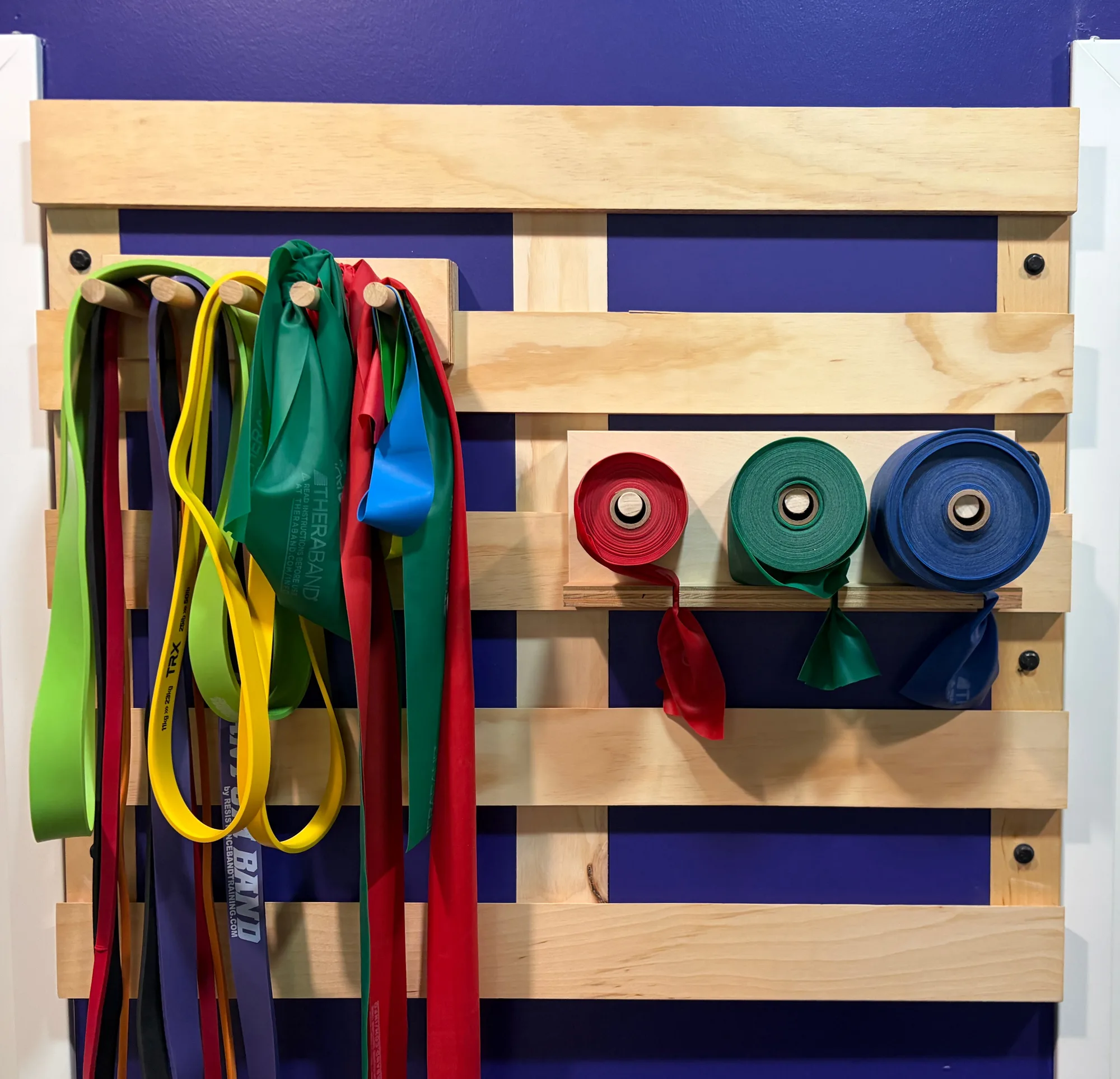 Wall-mounted plywood slat panel holding resistance bands on pegs and rolled therapy band spools